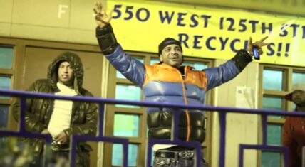 Jim Jones stands outside, wearing a puffer jacket, raising his arms, Harlem street scene.