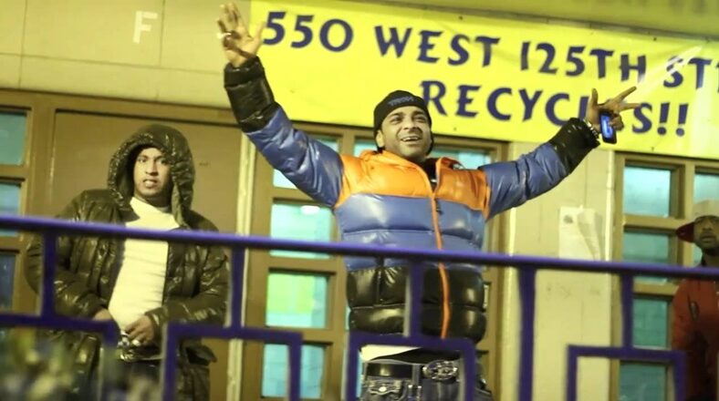 Jim Jones stands outside, wearing a puffer jacket, raising his arms, Harlem street scene.