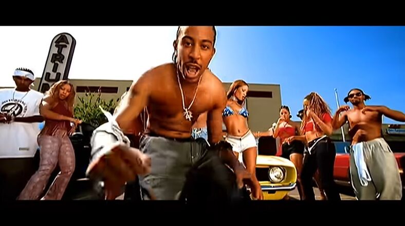 Ludacris performing shirtless in the "What's Your Fantasy" music video, surrounded by dancers and a vibrant party atmosphere.