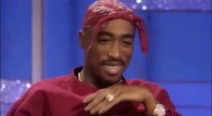 Tupac Shakur smiling during his second appearance on The Arsenio Hall Show in 1994, wearing a red bandana and a cross necklace, as he speaks candidly about his life and career.