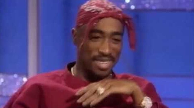 Tupac Shakur smiling during his second appearance on The Arsenio Hall Show in 1994, wearing a red bandana and a cross necklace, as he speaks candidly about his life and career.