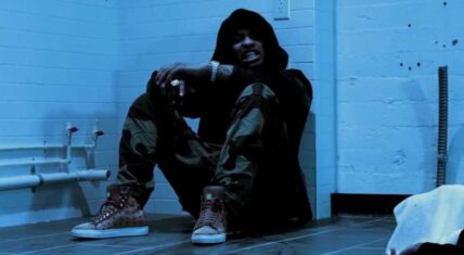 Waka Flocka Flame sitting against a wall in a dimly lit room wearing camo pants and a black hoodie.