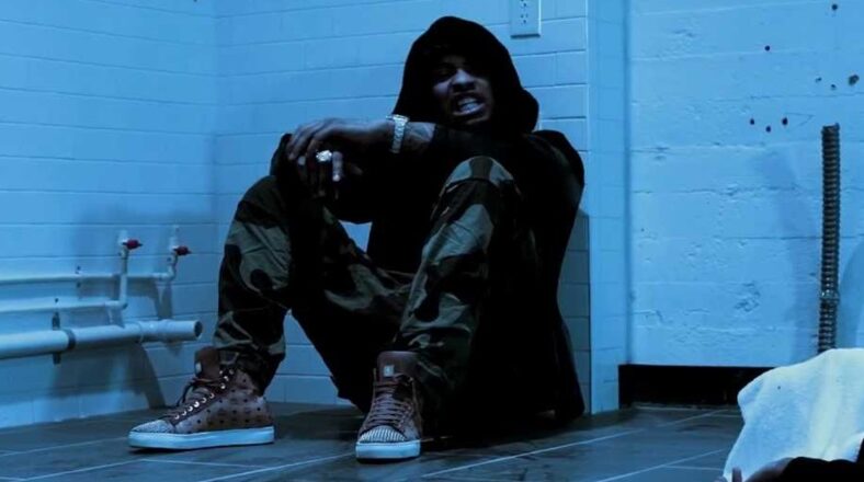 Waka Flocka Flame sitting against a wall in a dimly lit room wearing camo pants and a black hoodie.