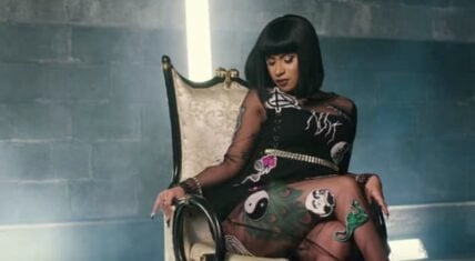 Cardi B sitting on a throne-like chair in the “Bodak Yellow” music video, wearing a sheer black dress adorned with colorful designs.