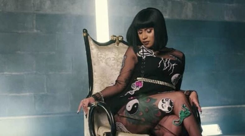 Cardi B sitting on a throne-like chair in the “Bodak Yellow” music video, wearing a sheer black dress adorned with colorful designs.