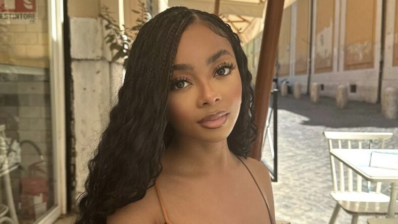 Skai Jackson sitting outdoors in a casual setting, with long, curly hair and a neutral-toned outfit, looking directly at the camera with a calm expression.