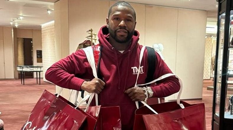 Floyd Mayweather holding luxury shopping bags, wearing TMT-branded clothing, showcasing his opulent lifestyle.