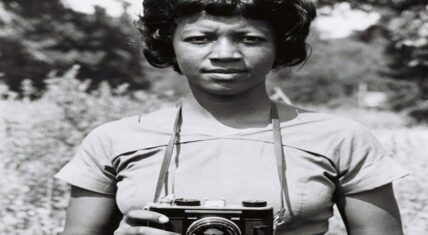 Photorealistic portrait of Doris Derby-inspired civil rights activist and photographer: a determined young Black woman in a simple 1960s dress holds a classic camera, standing amid tall grass and trees, symbolizing her decade-long documentation of the freedom struggle in Mississippi with SNCC (1962–1972).
