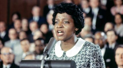 Photorealistic reimagination of Fannie Lou Hamer, the renowned civil rights activist and Mississippi Freedom Democratic Party delegate, testifies passionately before the Credentials Committee at the 1964 Democratic National Convention in Atlantic City. Seated at a long table with a large microphone, she speaks intently into the mic, hands clasped, wearing a light-colored collared blouse. In the background, blurred figures of convention attendees and officials stand beneath dramatic overhead lighting, capturing a pivotal moment of her historic challenge to segregated Southern delegations.