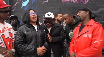 Supa Hot Fire (in black hoodie and blue sunglasses) facing off with Ray J (in red jacket) before their Brand Risk 14 fight, surrounded by a group of men in front of a graffiti wall.
