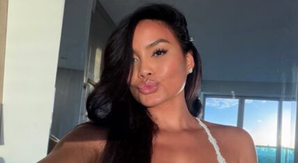 Daphne Joy posing for a selfie with long dark wavy hair, glossy lips, and a kissy face expression while wearing a white lace top