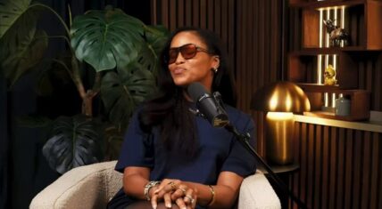 Eve seated in a studio armchair during her interview on The Romesh Ranganathan Show, wearing sunglasses and a navy top while speaking into a microphone, with a monstera plant and illuminated shelves visible in the background.