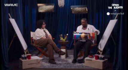 Funny Marco sits in a director's chair holding a colorful poster box while speaking with Raven-Symoné, who is seated opposite him in a studio interview setup featuring blue curtains, easels with blank canvases, and art supplies.