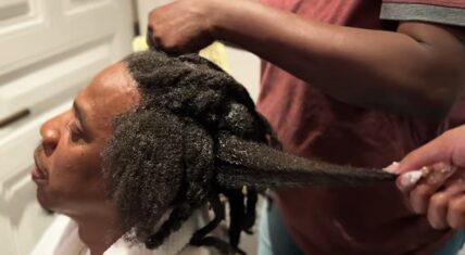 Close-up of Jay-Z during his CÉCRED hair transformation, with hands applying product and stretching a long, saturated loc as part of the multi-day locs-to-afro process.