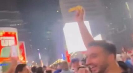 Knicks fan with a big smile holding a yellow banana high in the air amid a dense nighttime crowd celebrating in New York City streets, with bright billboards glowing in the background.