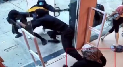 CCTV footage of a security guard physically blocking two suspects attempting to enter a New York store during a robbery attempt. A woman in pink stands nearby outside.