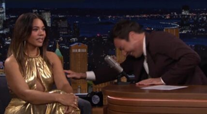 Regina Hall in a striking gold dress smiles while Jimmy Fallon laughs and leans across the desk on The Tonight Show set with a New York City skyline backdrop.