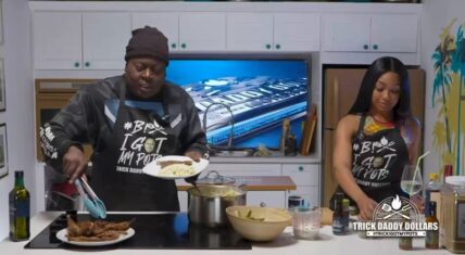 Trick Daddy wearing a black beanie and apron serves a plate of steak and mashed potatoes with tongs while standing next to Yung Miami in his "B! I Got My Pots" kitchen set. Yung Miami, also in a branded apron, holds a wine glass and watches.
