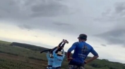 Workers in blue shirts lift a woman in a helmet and harness into a horizontal launch position at the edge of Ponte do Esqueleto bridge during the fatal incident in Brazil, as a man in a blue shirt and cap stands watching with hands on hips; rural landscape under overcast sky.