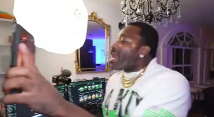 Adrien Broner gets emotional checking his phone in a luxury room during his Kick streaming comeback, gold chains and white t-shirt visible.