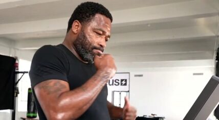Adrien Broner sweaty and focused on a treadmill in the gym during his 45-day fitness challenge, wearing a black t-shirt with visible arm tattoos, giving a thumbs-up gesture while catching his breath.