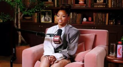 Chlöe Bailey seated in a pink velvet armchair during her Call Her Daddy podcast interview, wearing a striped shirt under a gray blazer with hair in a high bun, smiling while speaking into a microphone against a backdrop of bookshelves.