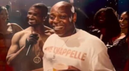 Dave Chappelle smiling widely on stage next to shirtless Usher during a performance of 