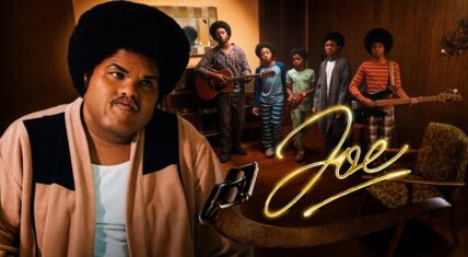 Druski as Joe Jackson in the foreground wearing a large afro and beige jacket, staring seriously at the camera, with the Jackson 5 brothers holding guitars in a 1970s living room behind him. A leather belt rests on the table beside a glowing yellow neon 