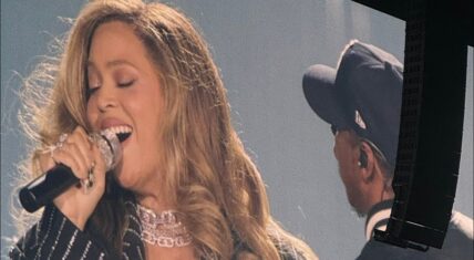 Beyoncé performing on stage, singing passionately into a microphone with her eyes closed and mouth open, wearing a sparkling pinstripe outfit, diamond choker necklace, and rings, with long wavy blonde hair; Jay-Z is visible in the background wearing a dark baseball cap and in-ear monitor.