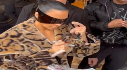 Kim Kardashian, wearing oversized dark shield sunglasses and a leopard-print fur coat, leans forward to sign an autograph with a marker while surrounded by security and fans.