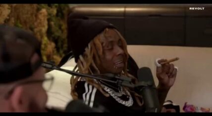 Lil Wayne speaking during an interview on DJ Drama's Gangsta Grillz Podcast, wearing a black beanie and track jacket while holding a cigar, with a microphone in front and the interviewer partially visible in the foreground (Revolt TV).