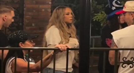 Mariah Carey standing patiently in a nighttime queue line at Disneyland Resort, wearing a light-colored top and blue jeans with her hands resting on a metal railing, surrounded by other park guests including a woman in a sparkly cap and a person in a sorcerer hat.