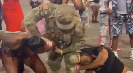 National Guard soldier in camouflage intervenes in a street fight on Beale Street pavement, physically separating a woman with long dark hair and another woman in a black top as bystanders watch and record.