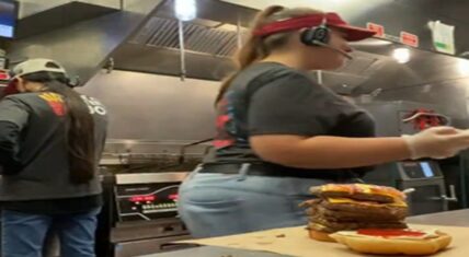 Wendy's female employee wearing headset and gloves stands at the kitchen counter beside a massive multi-patty stacked burger with separate bottom bun, another worker visible in the background.